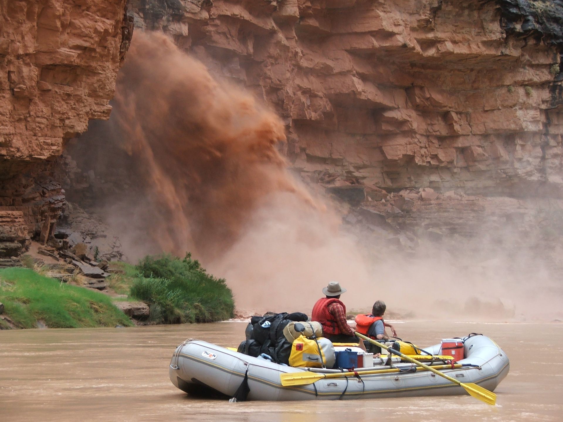 Rafting on a muddy river, below a waterfall of reddish dust. Two people in the raft. Red rock canyon.