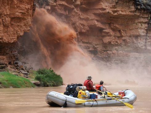 Rafting on a muddy river beneath a sand-laden waterfall with rocky canyon walls.