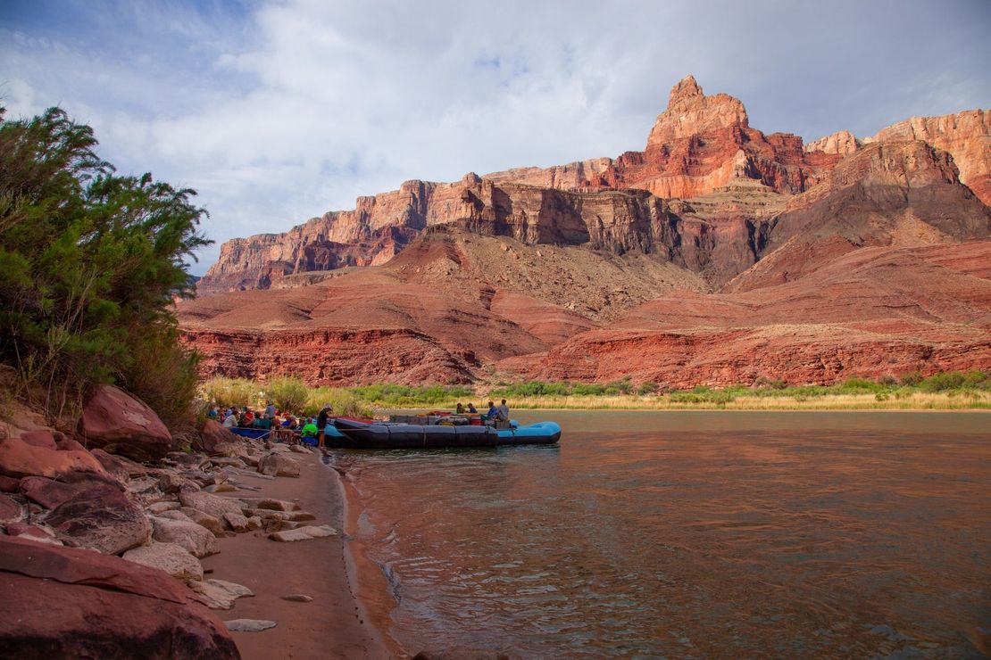Raft on riverbank at base of large red rock cliffs. Cloudy sky.