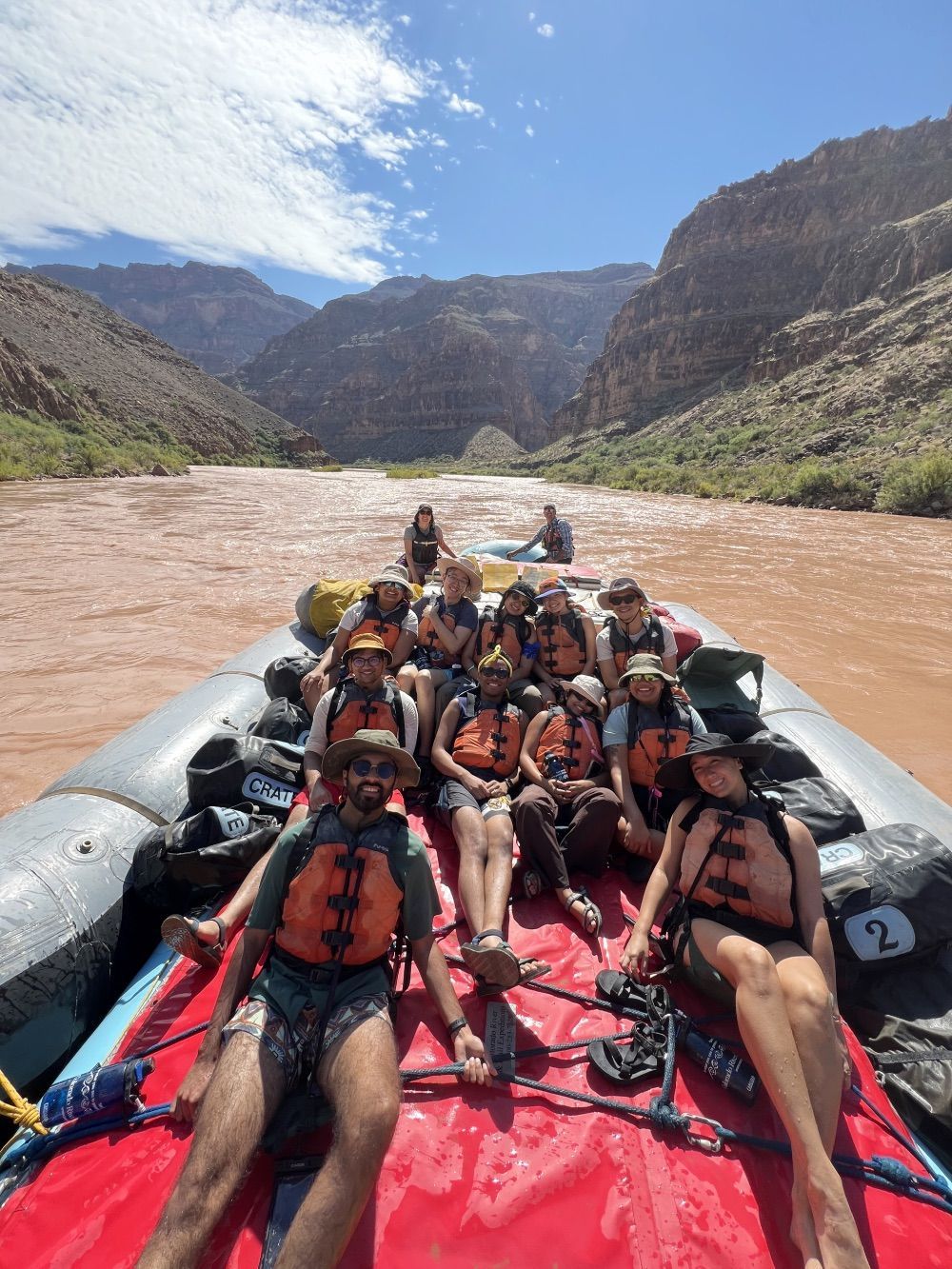 Group of people on a raft in a red river between canyon walls under a blue sky.
