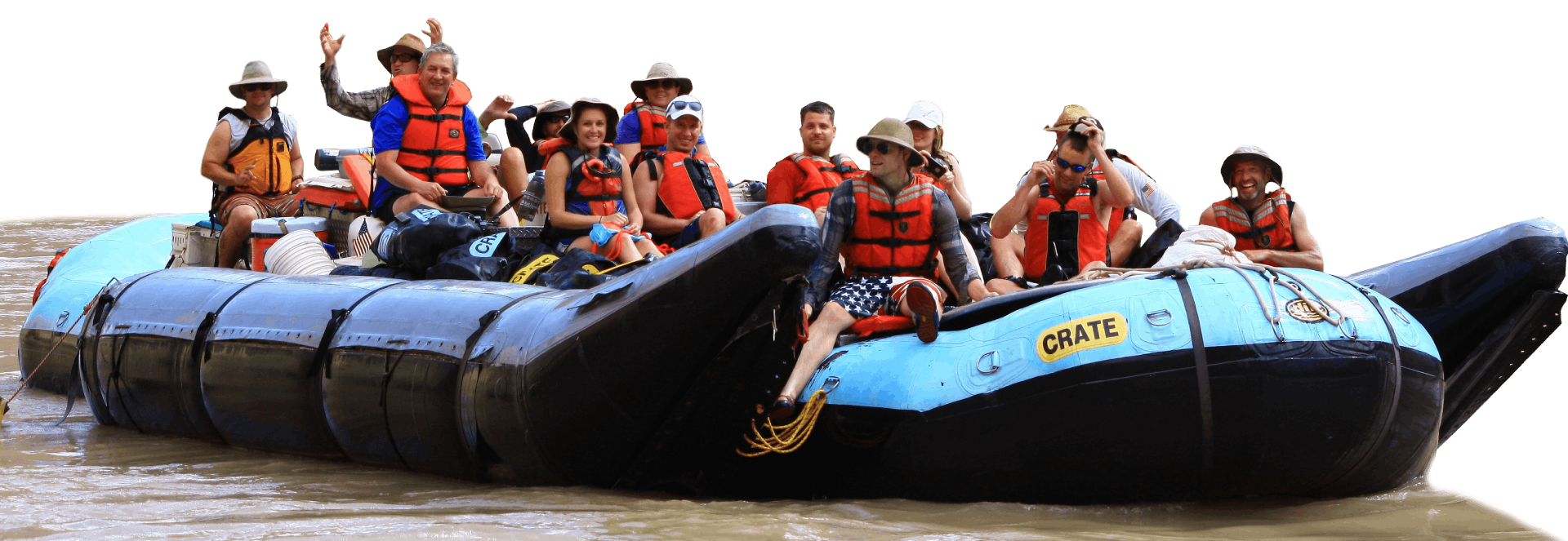 People in life vests on a raft in a river, with some waving.
