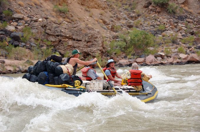 Rafting on a river; four people paddling through rapids. White water, rocky cliffs in background.