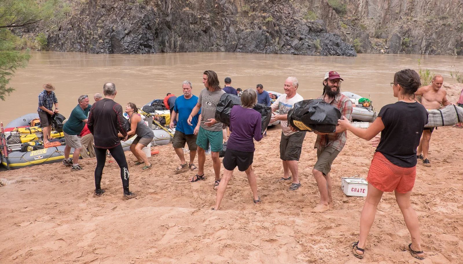 People on a sandy riverbank holding and carrying gear near rafts. Brown water and trees in the background.