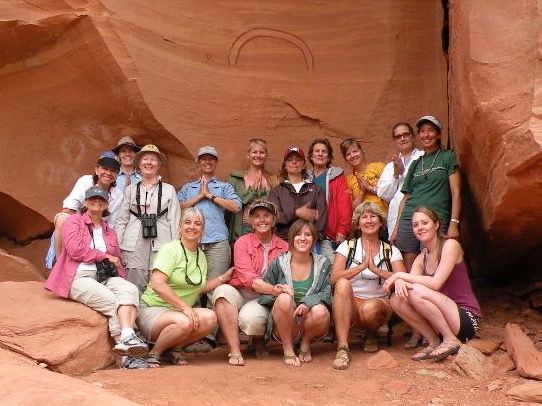 Group of people posing by a sandstone wall, some in yoga poses. Red-orange rock, bright daylight.