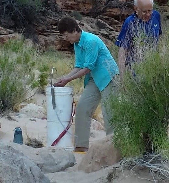 Woman in turquoise shirt washing hands over a bucket; man in blue shirt watches nearby, outdoor setting.