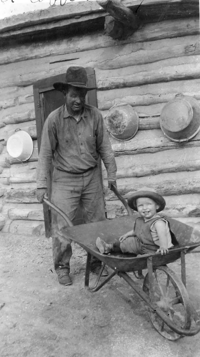 Man pushing wheelbarrow with child in front of log cabin.