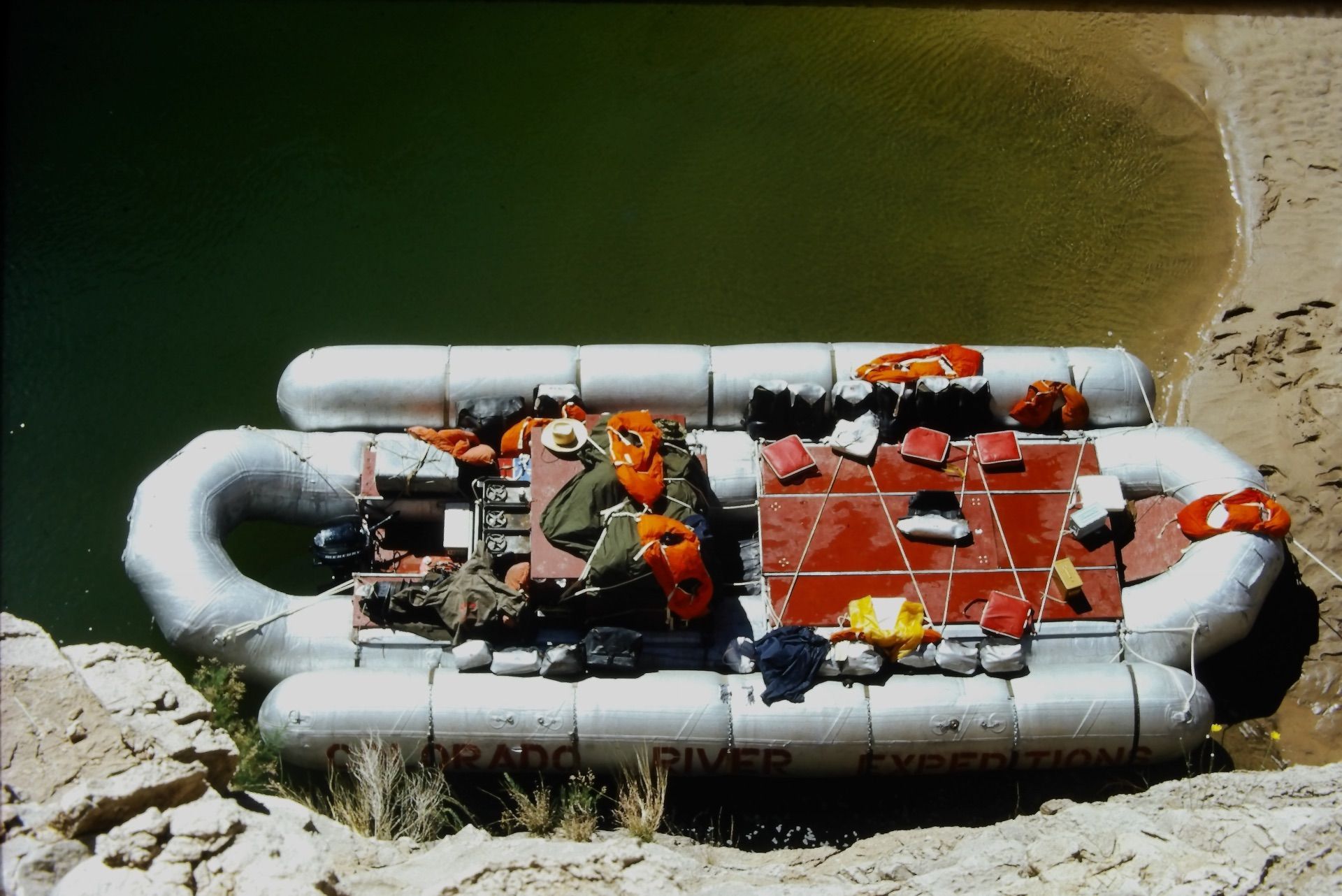 Raft on the shore of a green river. Several life vests and gear are on the raft.