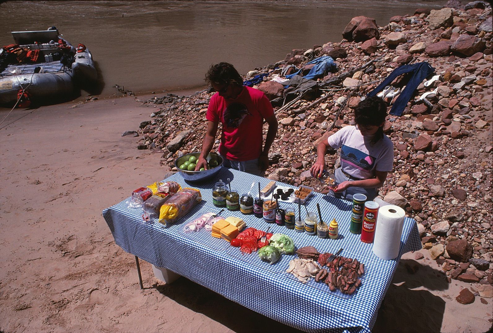 Two people prepare food on a picnic table on a sandy riverbank; a raft is nearby.