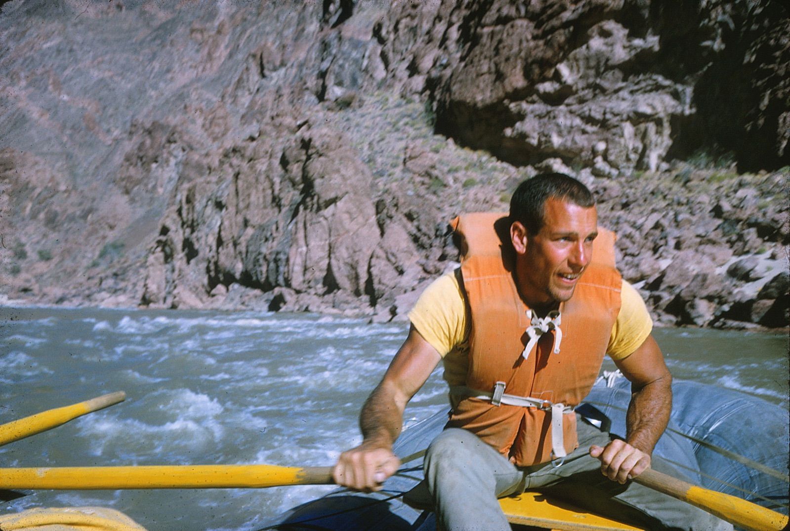 Man in a life vest rows a raft on a river, with canyon walls in the background.