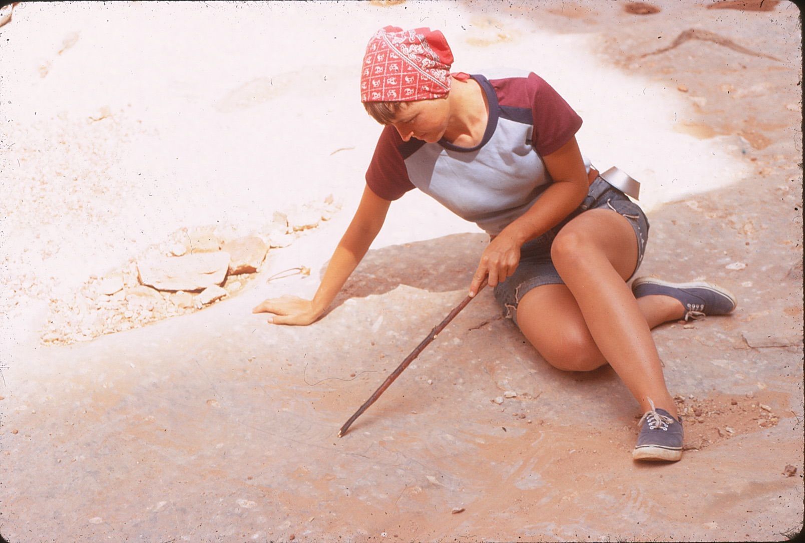 Woman kneeling outdoors, pointing with a stick at a light-colored surface; wearing a red bandana, blue shirt, denim shorts.
