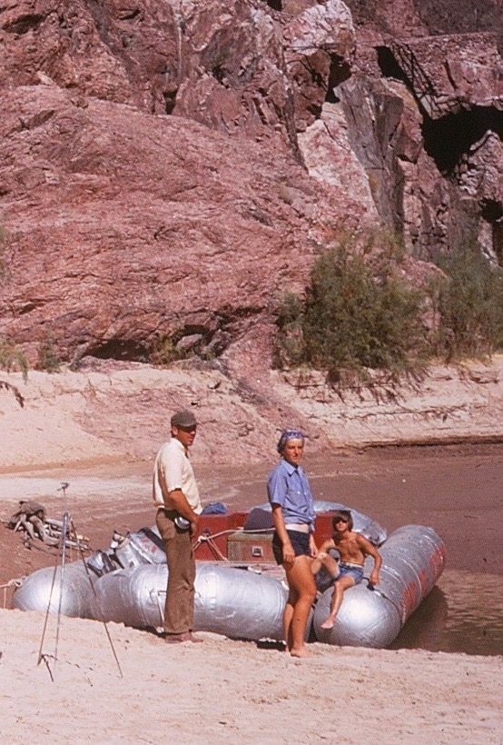 People and raft on a sandy riverbank, with reddish-brown rock cliff in the background.