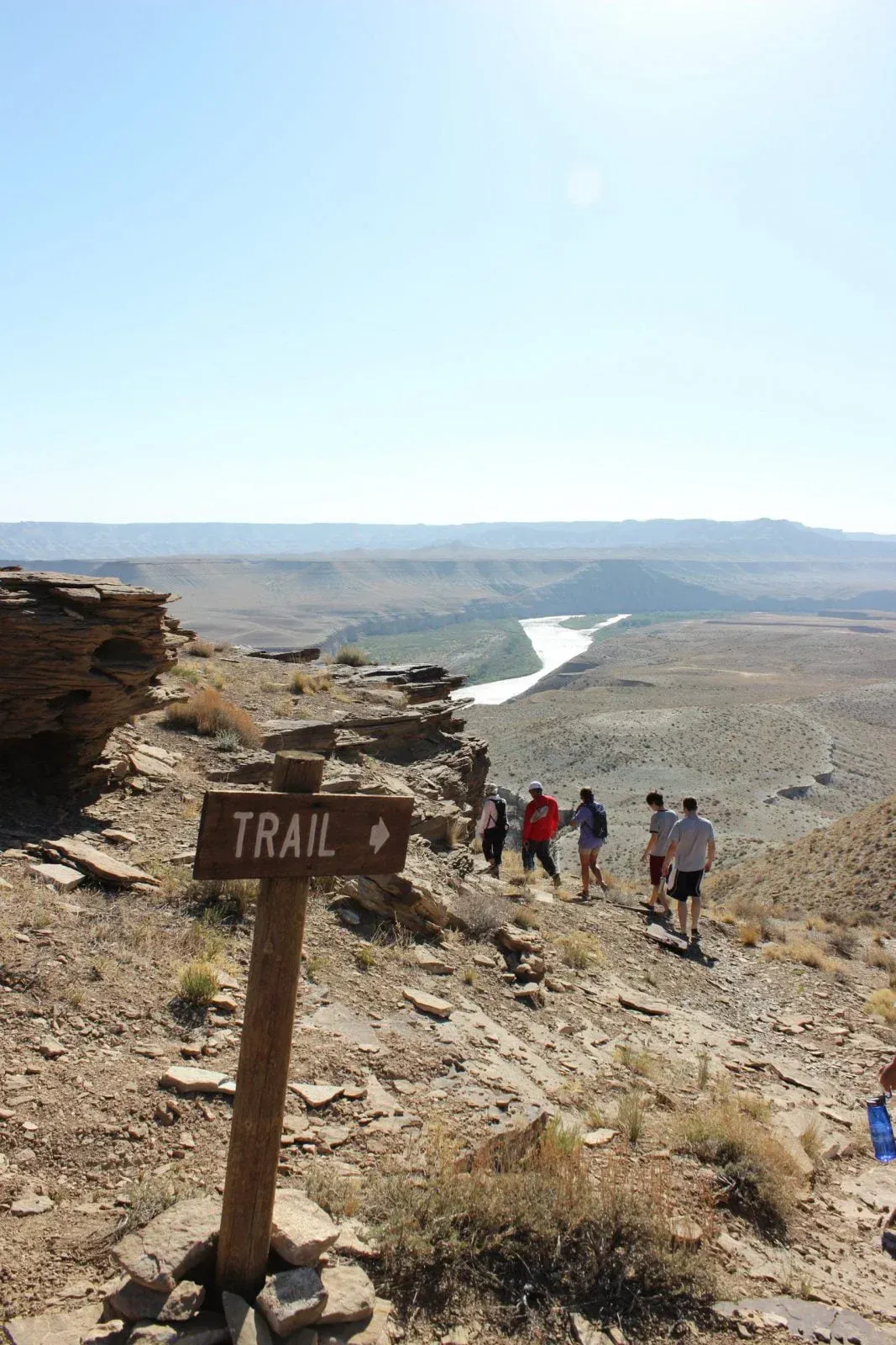 Trail sign points toward a canyon with people hiking, river visible in the distance under a blue sky.