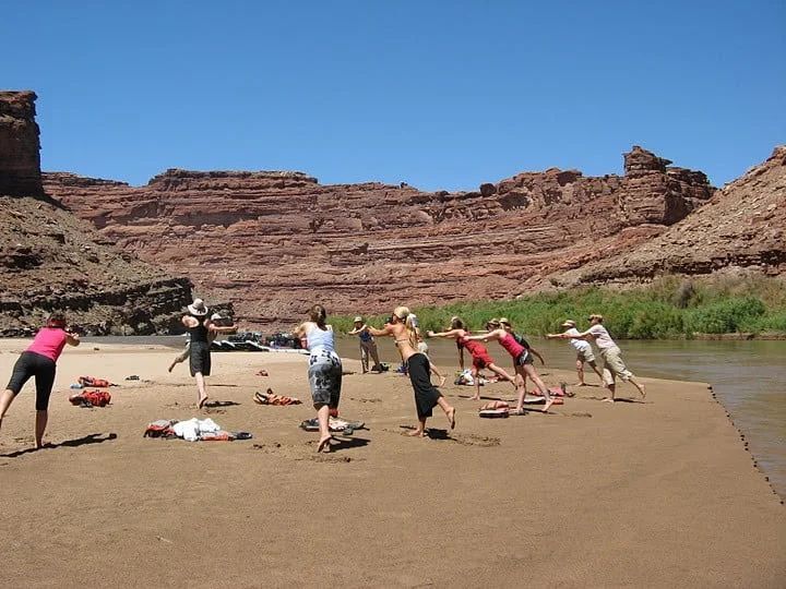 Group doing exercises on a sandy riverbank with red rock cliffs in the background under a blue sky.