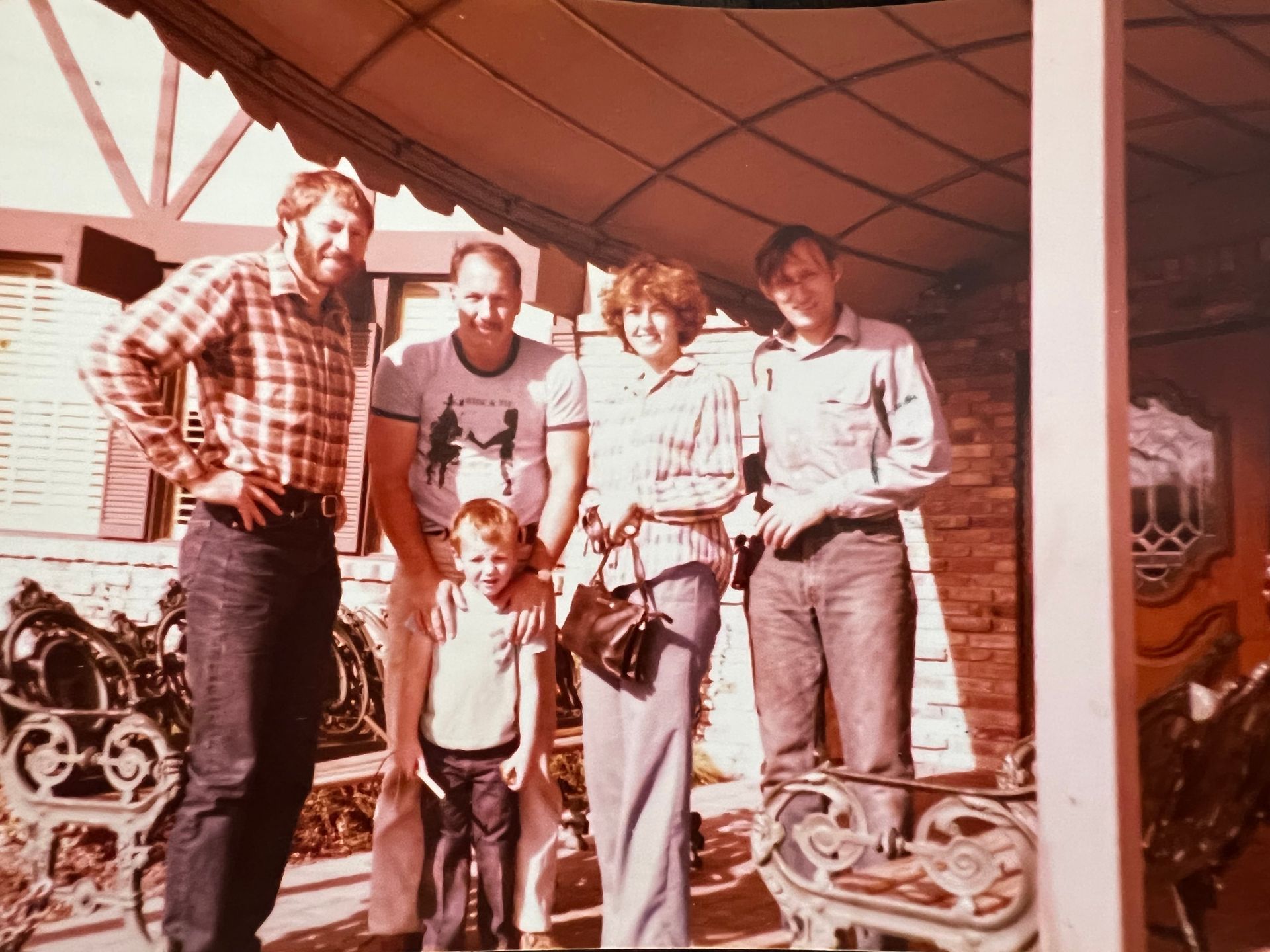 Group of five people posing outdoors, under a roof. People smiling, some with arms around each other. Vintage photo.