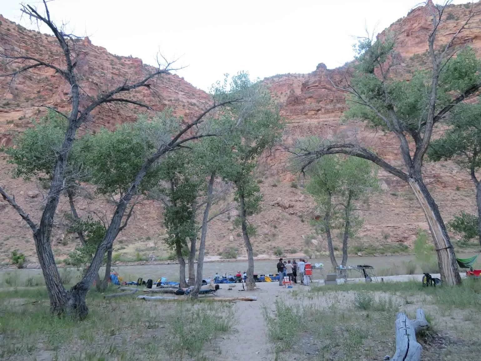 Campground with trees, group of people, tents, and river, set against red rock canyon walls.
