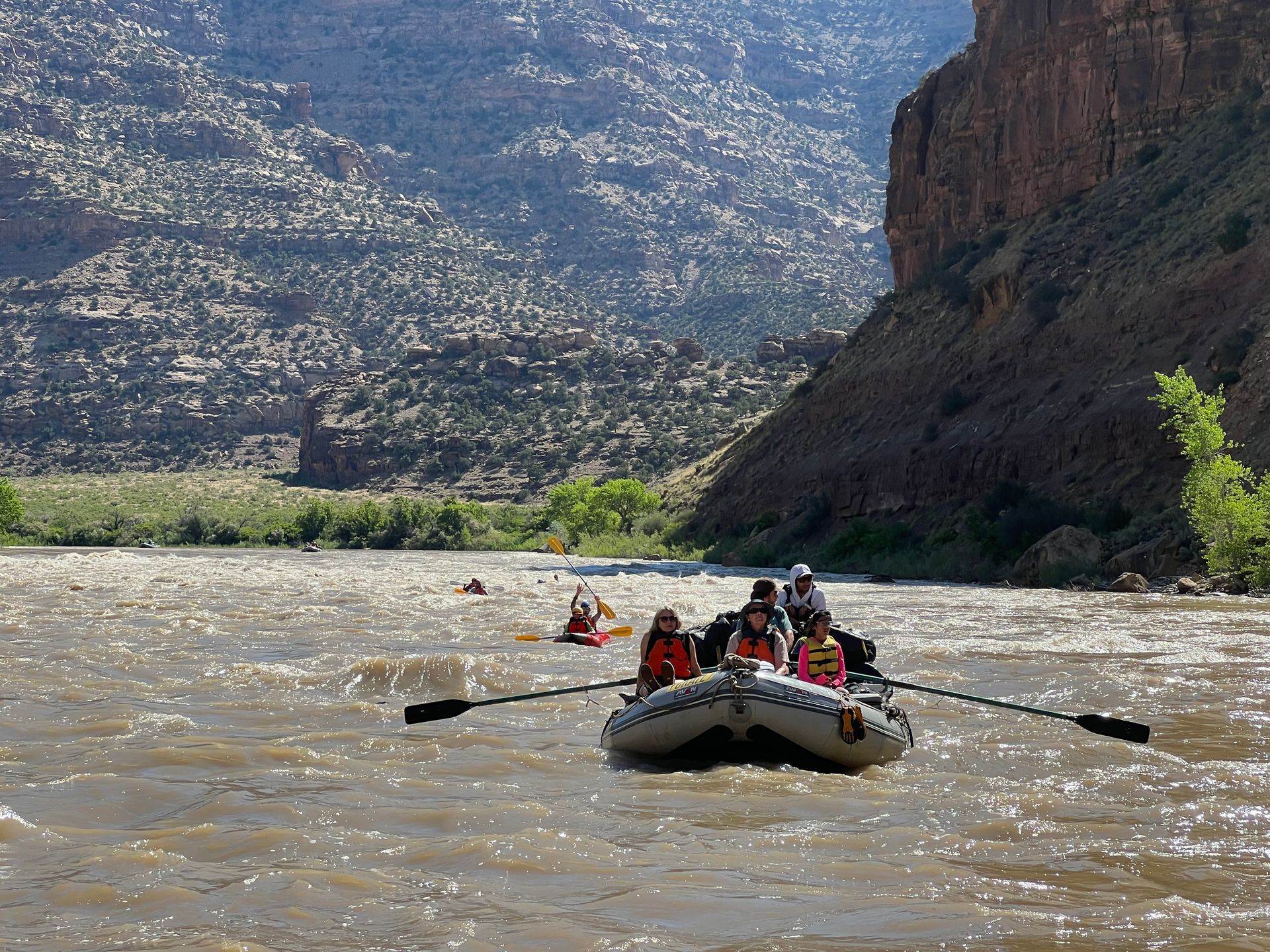 Rafting on a brown river, with people paddling, surrounded by canyons and hills.