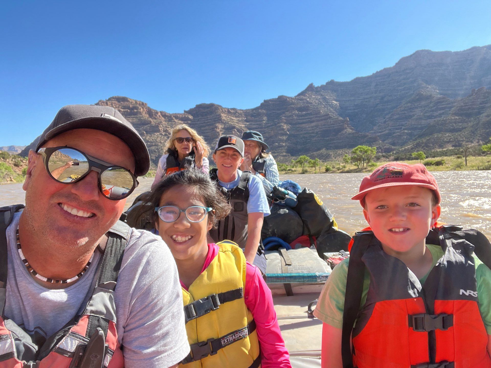 Group on a raft smiles for a selfie on a river with rocky, tan canyon walls in the background.
