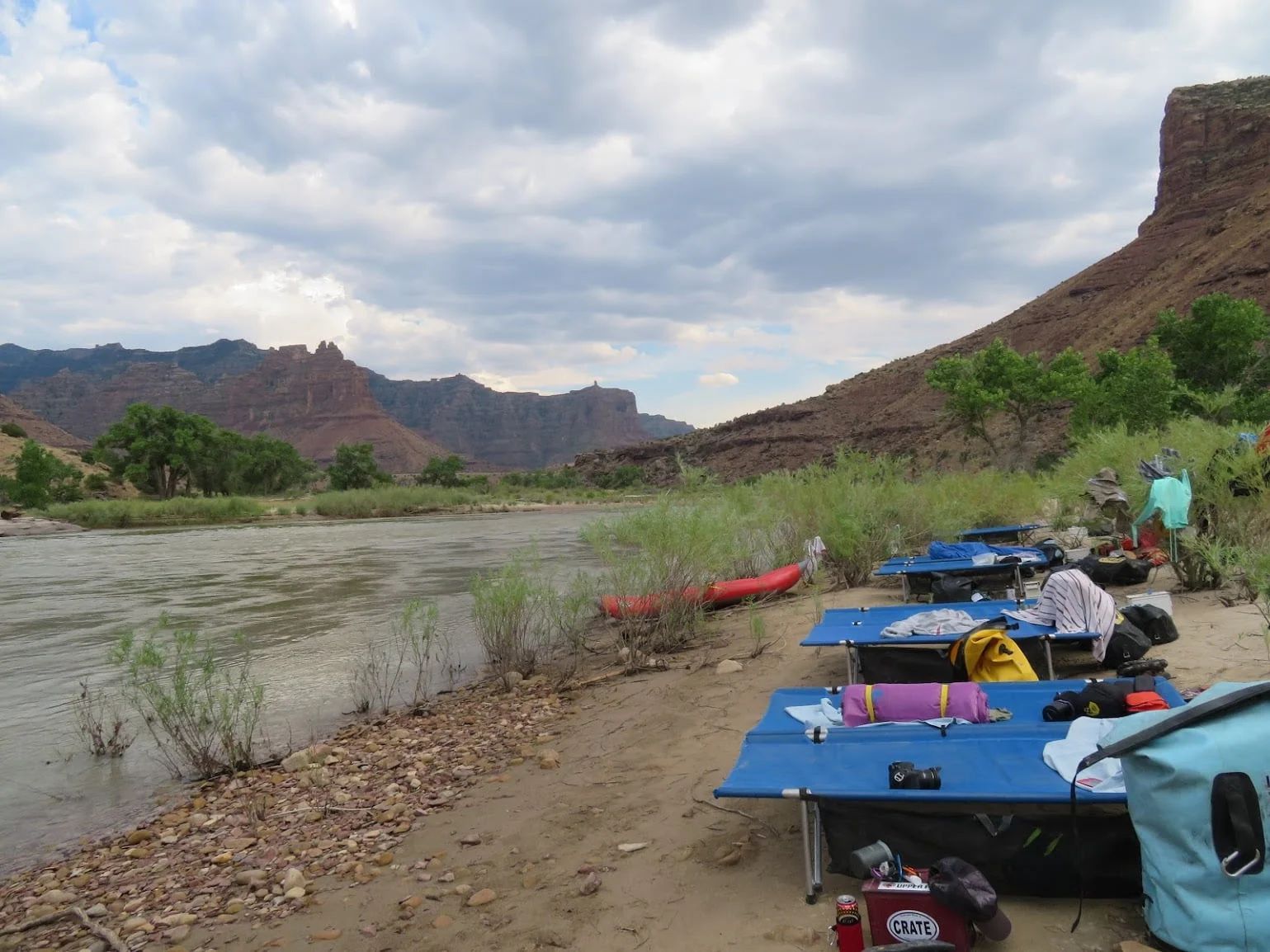 Riverbank campsite with blue cots, kayaks, and gear. Red rock cliffs and cloudy sky in background.