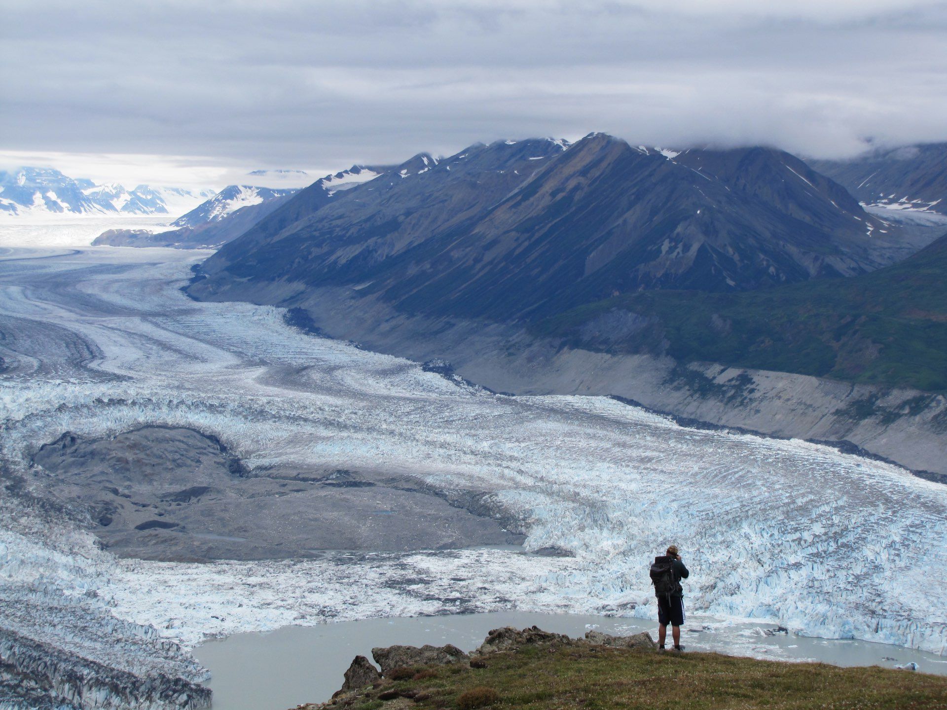 Man standing on cliff overlooking a glacier and mountains under a cloudy sky.