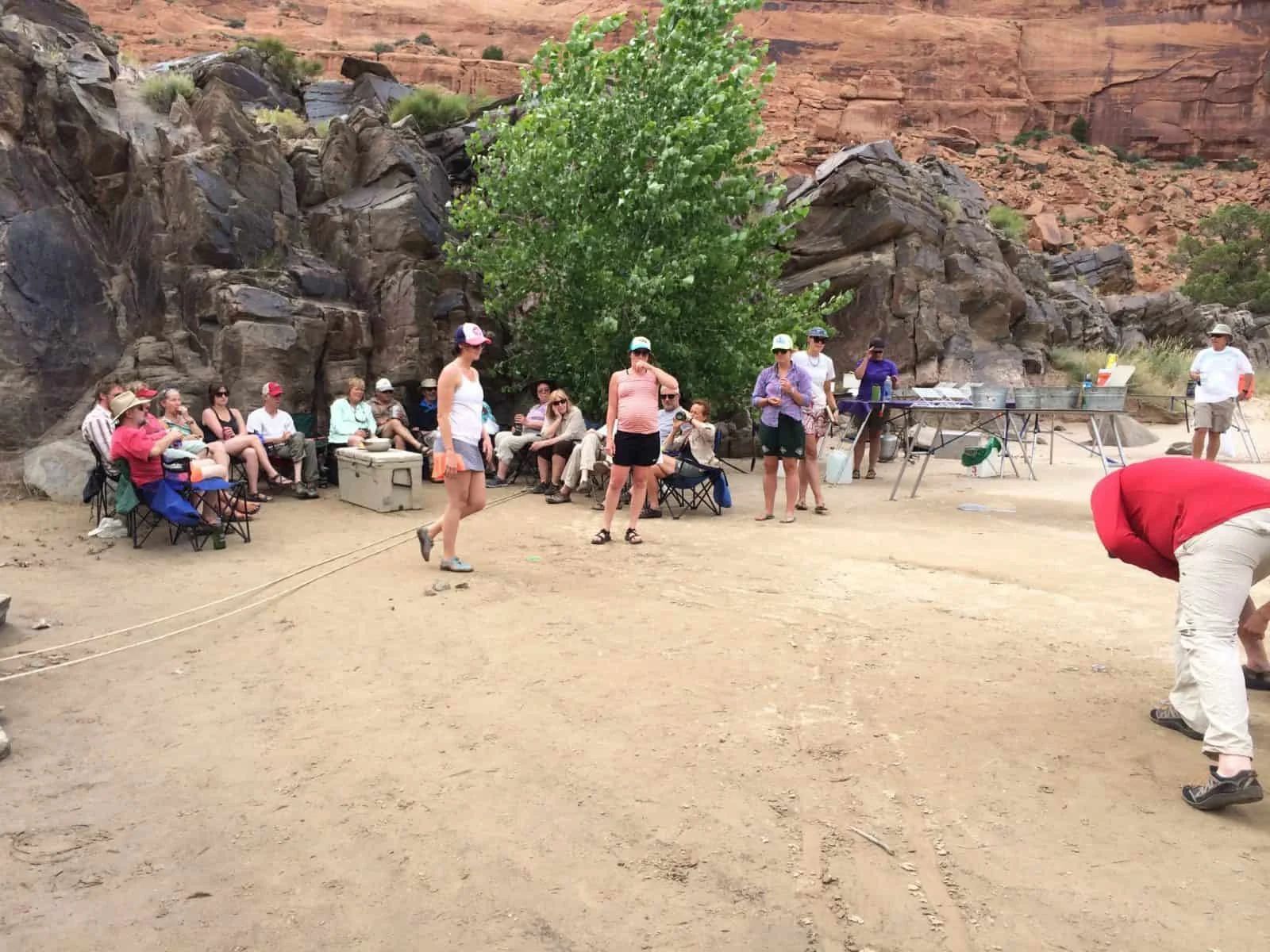Group of people socializing and playing games on a sandy terrain near rocks and trees.