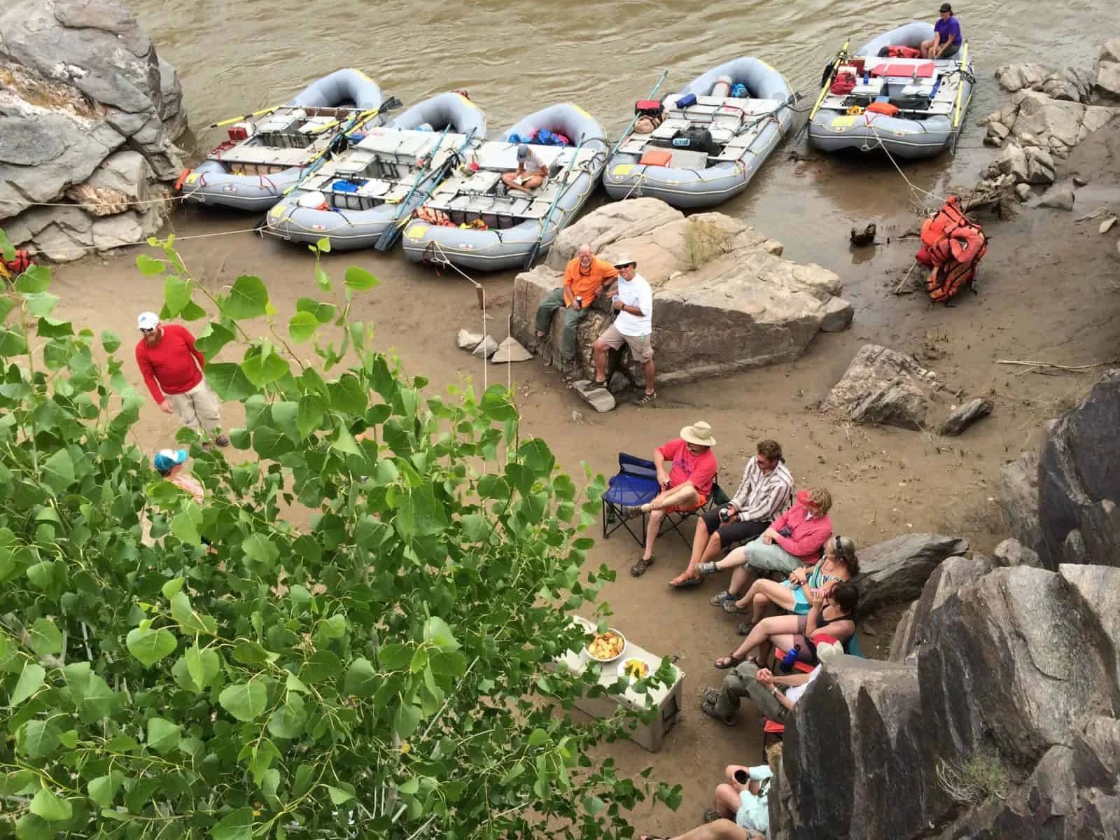 Rafts pulled ashore, people seated near river, watching. One man plays ball, others relax in the shade.