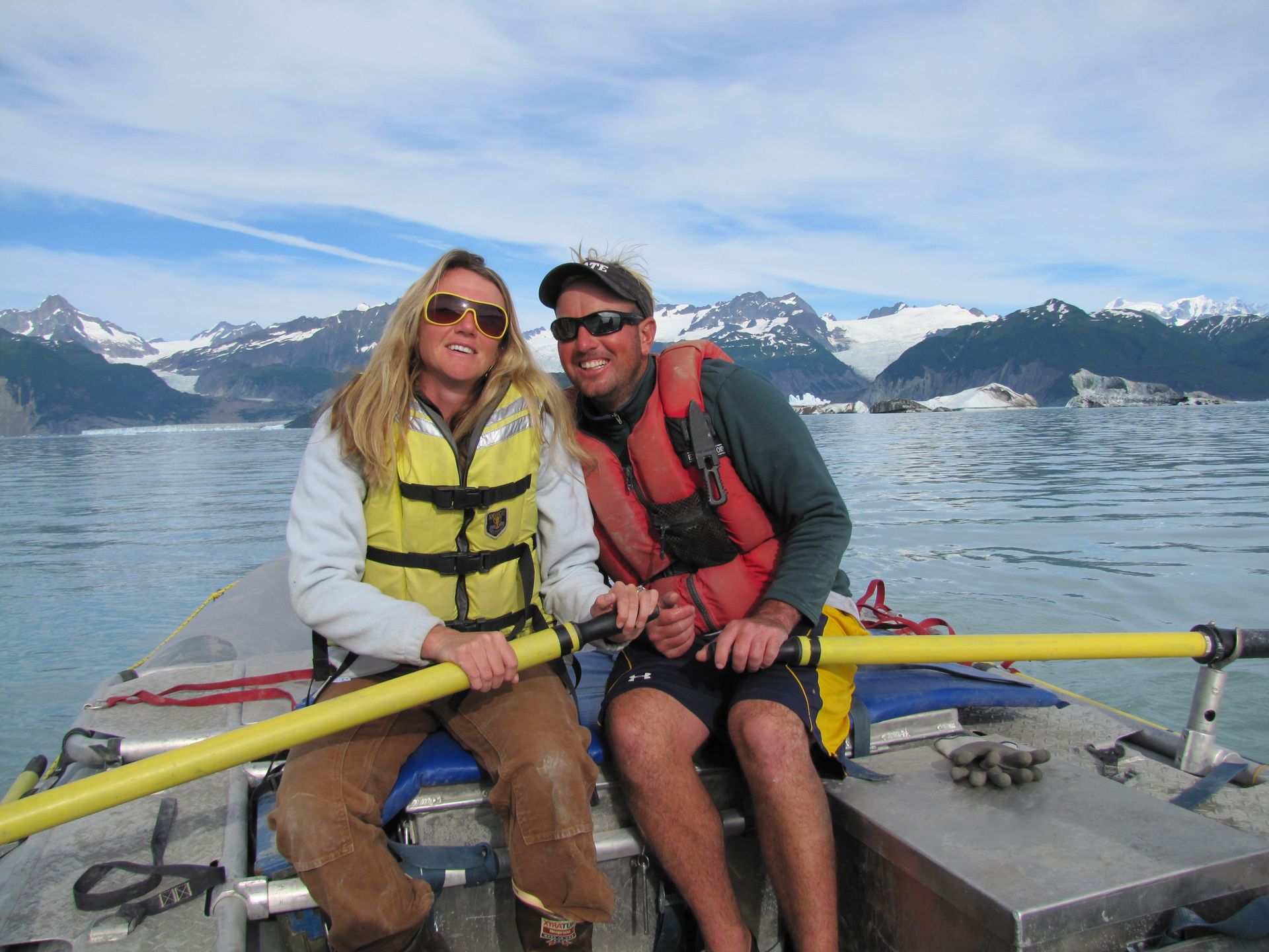 Two people in life vests on a boat with glacial mountains in the background.