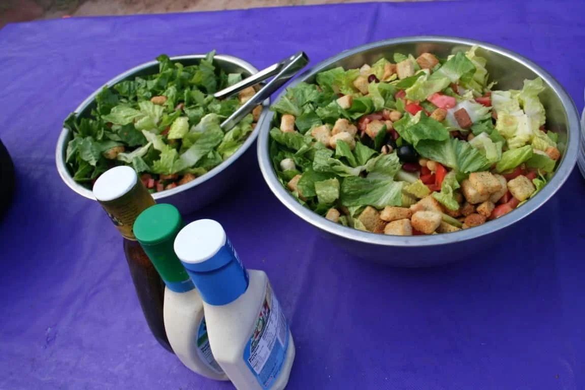 Two metal bowls of salad with dressing bottles on a purple tablecloth.
