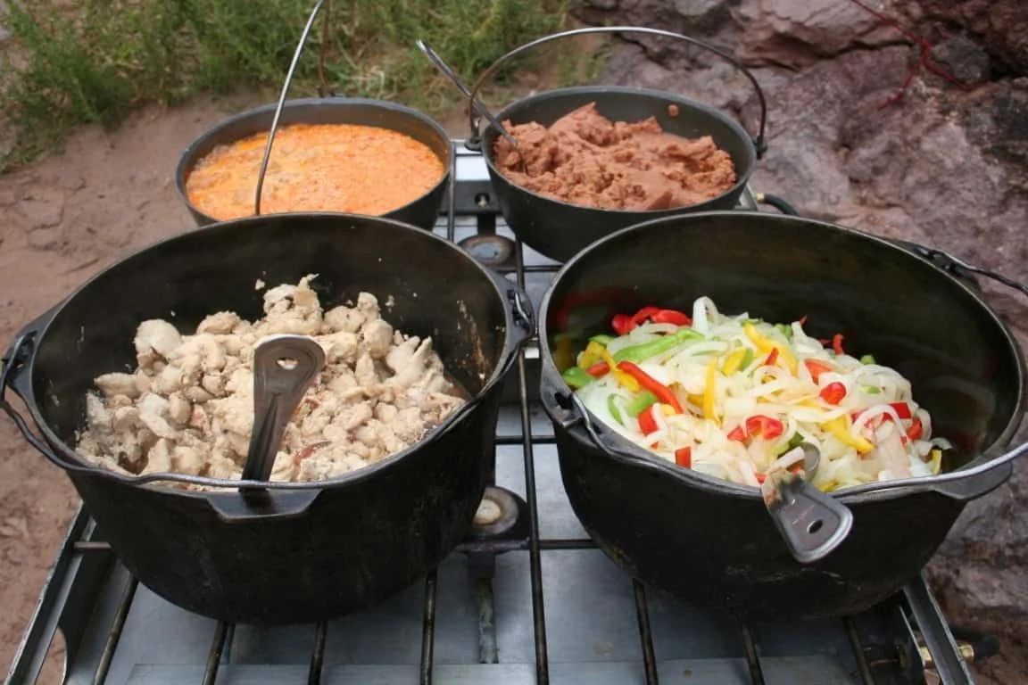 Four Dutch ovens on a camping stove; cooked chicken, beans, peppers, and cornbread are visible.