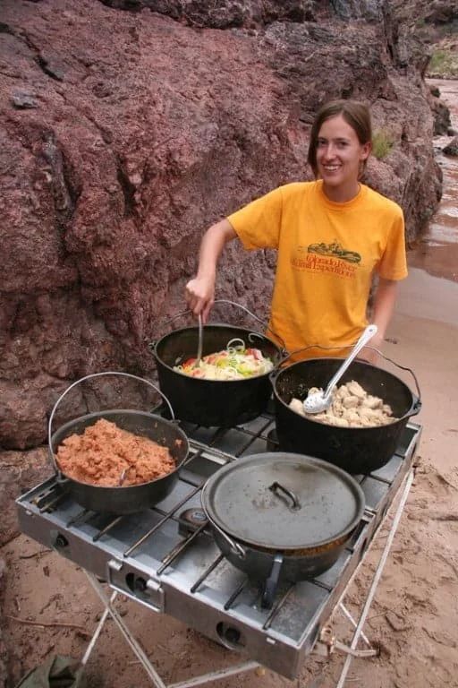 Woman cooking in Dutch ovens outdoors. She stirs food in one pot. Other pots sit on a camping stove.