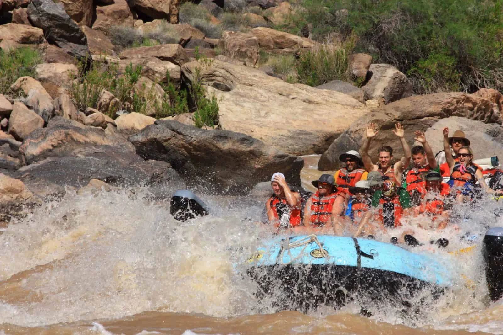 Rafting down a river, splashing through rapids. People in life vests, raising arms near rocky banks.