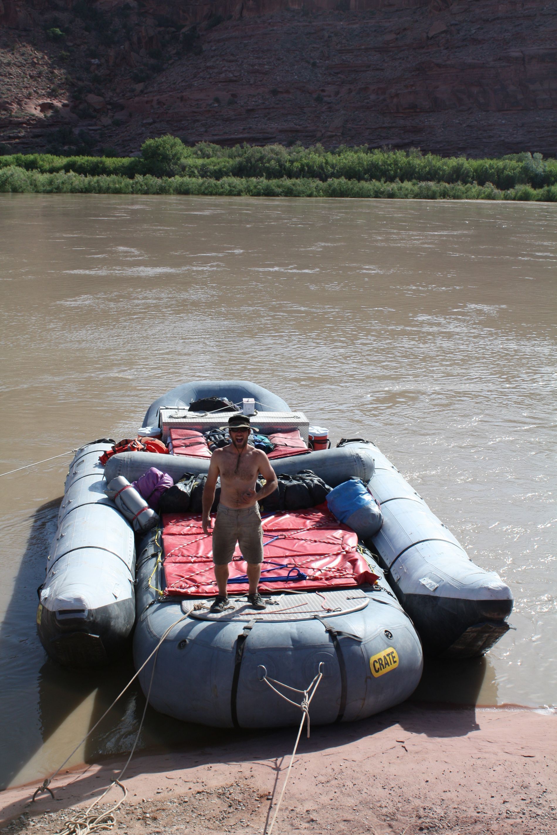 Man standing on a loaded raft on a muddy river, red rock cliffs in the background.