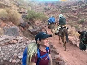 Woman hiker smiles at the camera on a rocky trail as riders on mules pass by.