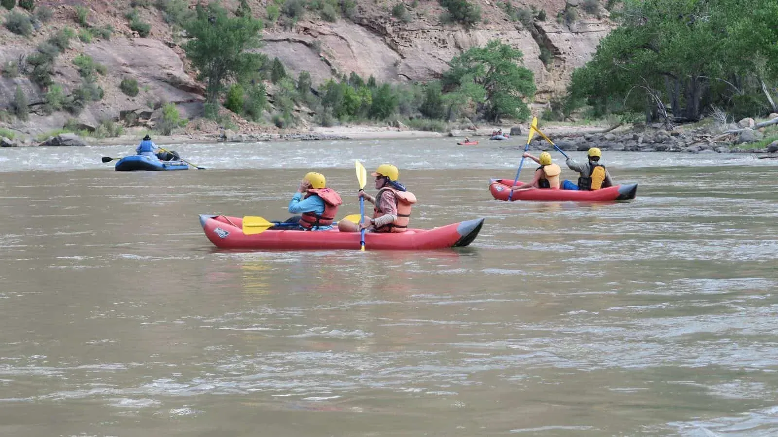 Kayakers in red boats with yellow helmets navigate a muddy river.  A larger blue raft is also on the water.