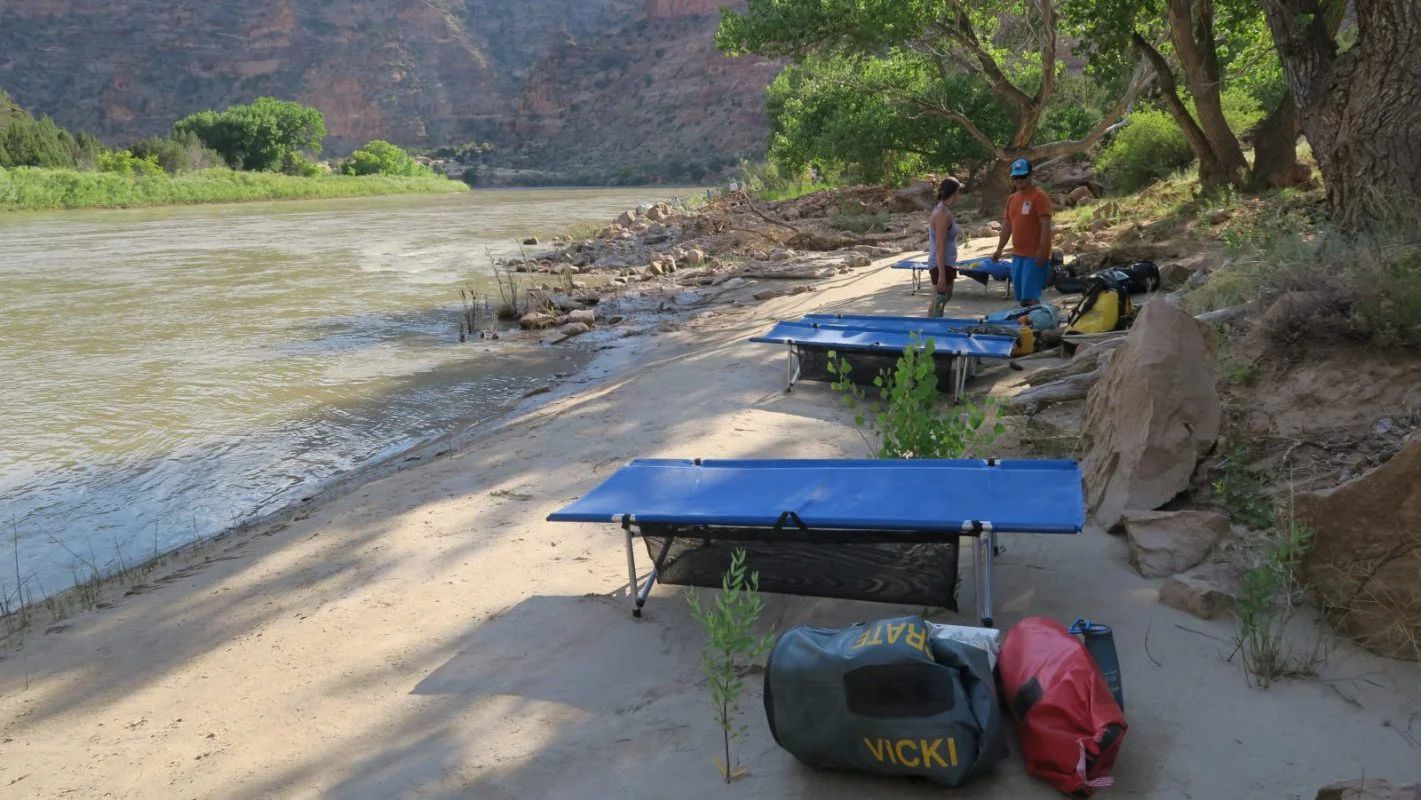 Two blue cots on a sandy riverbank, gear bags in front. Person standing near cots, trees in background.