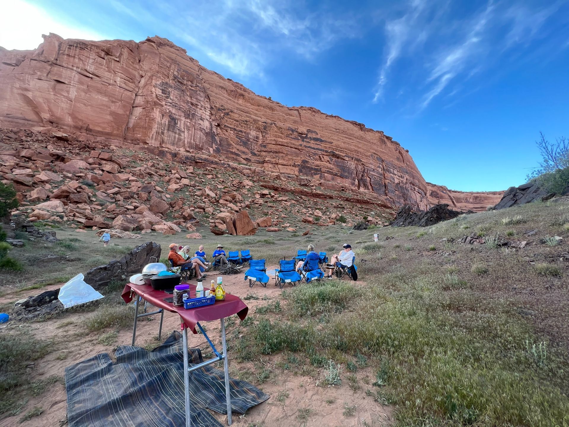 People in camp chairs near a red rock cliff, gathered around a table with supplies on a grassy area. Blue sky.