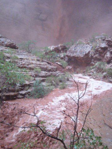 Flooded canyon waterway; red-brown water flows through a rocky channel, with greenery on the banks.
