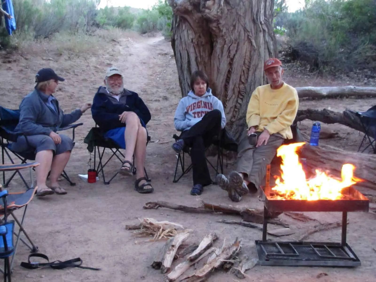 Four people sit in folding chairs around a campfire.