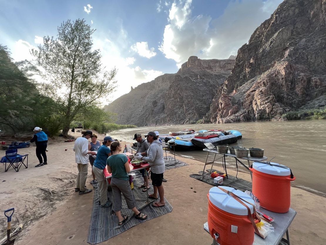 People preparing food at a riverside campsite with rafts. Mountains and sky in the background.