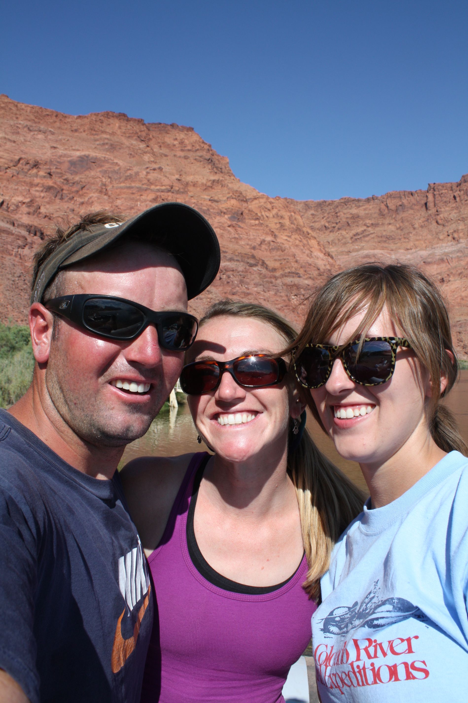 Three people smiling, wearing sunglasses, in front of a red rock canyon and blue sky.