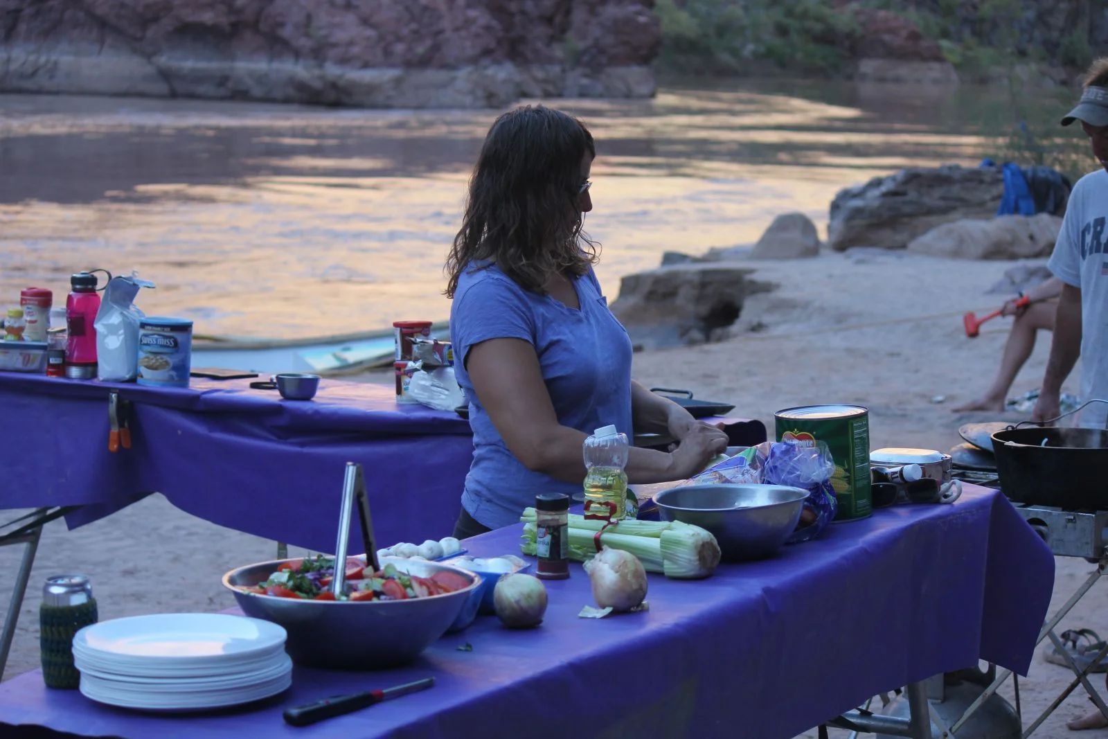Woman preparing food on a purple-covered table at a riverside campsite. Salad, vegetables, and condiments visible.