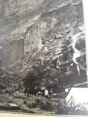 Rock formation with a waterfall and a group of people and horses gathered below it.