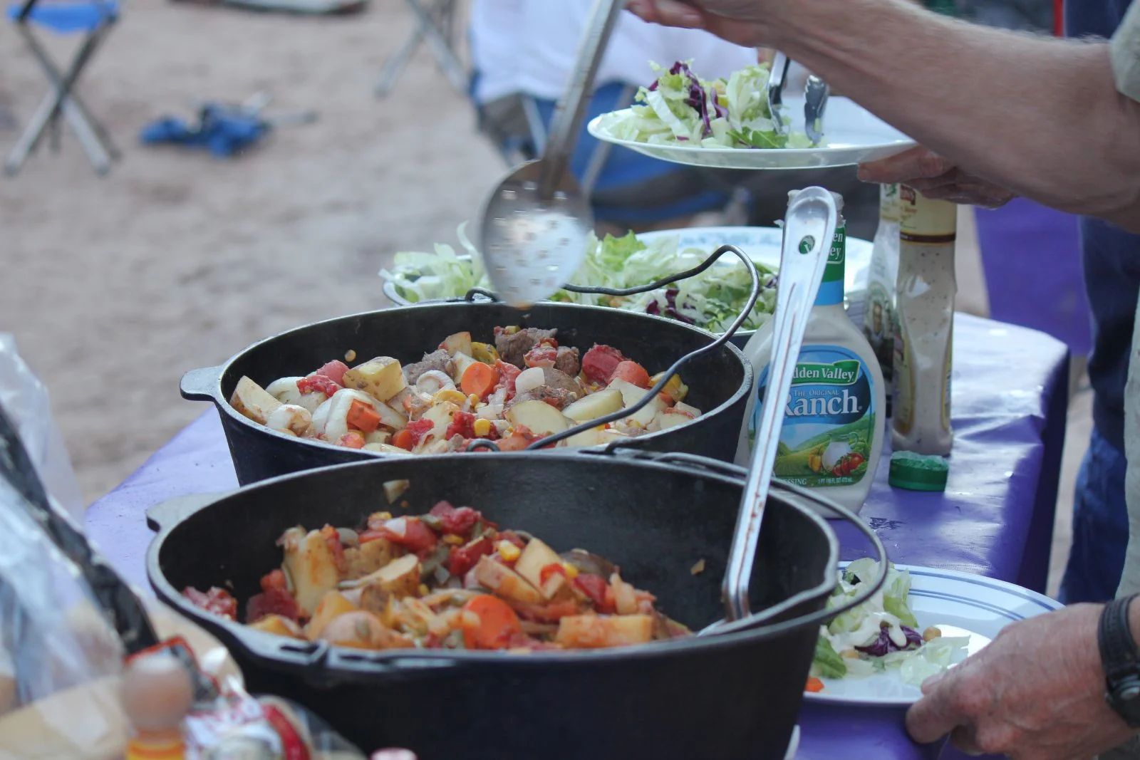 People serving food from Dutch ovens at an outdoor gathering. Salad on plates and a bottle of ranch dressing visible.