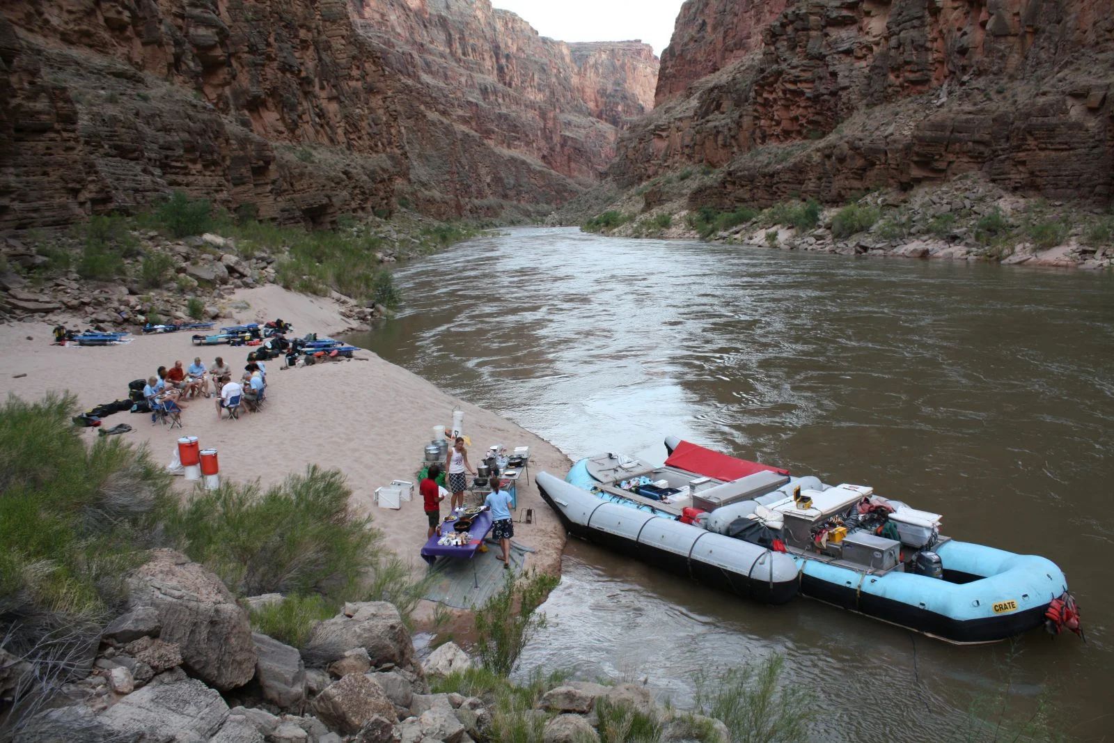 A large raft pulled up to a sandy riverbank, people unloading gear in a canyon.