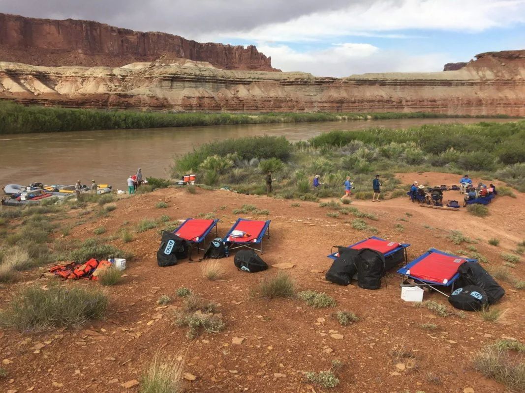 Rafting camp setup on a red-dirt riverbank, with rafts, gear, and people near a river and cliffs.