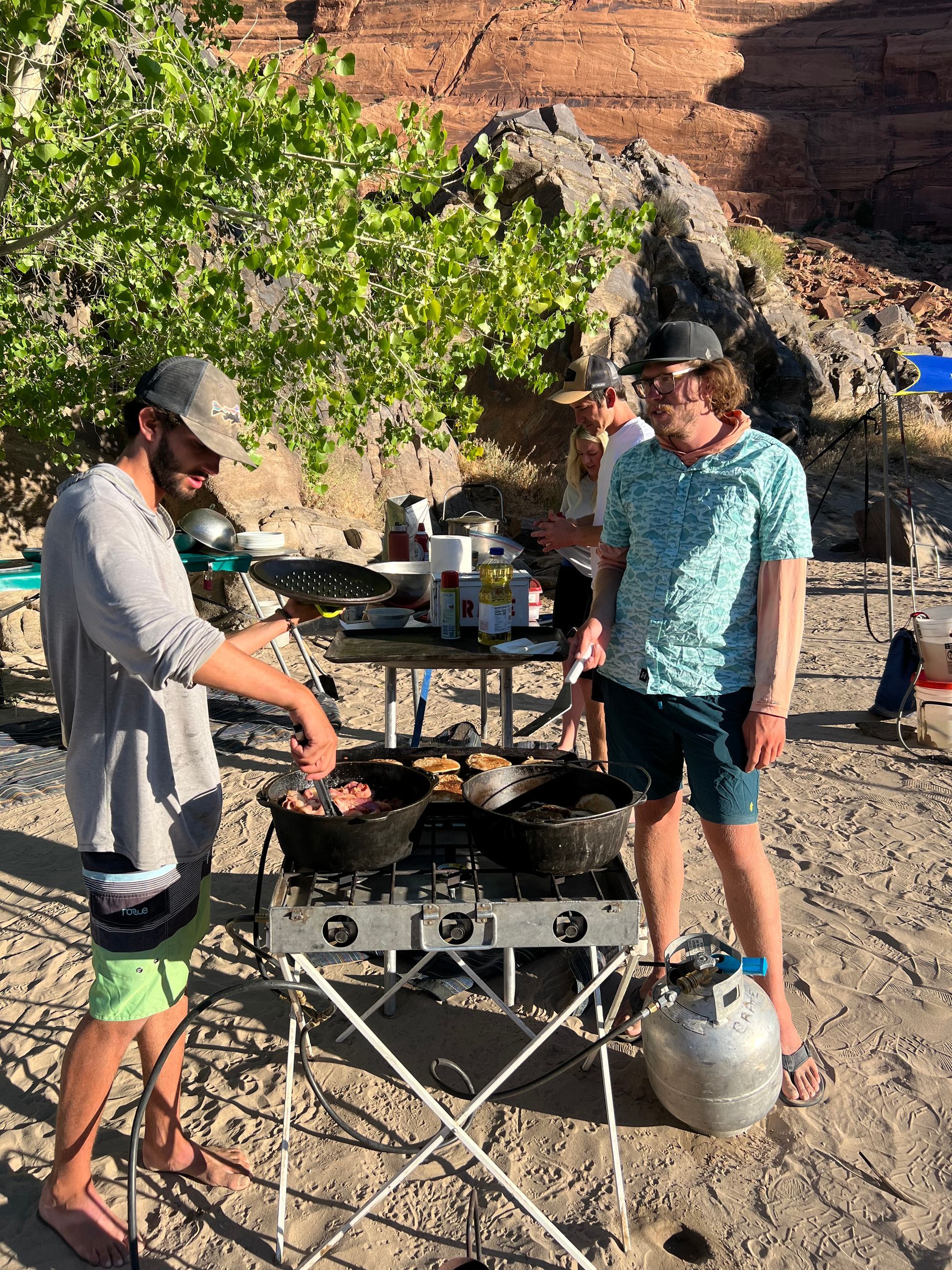 People cooking food outdoors on a portable stove, near a river with red rock cliffs and trees.