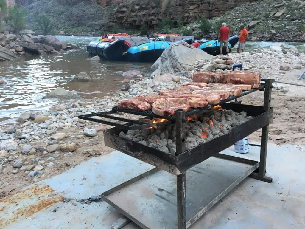 Steaks grilling on a metal grate near a river with rafts and people in the background.