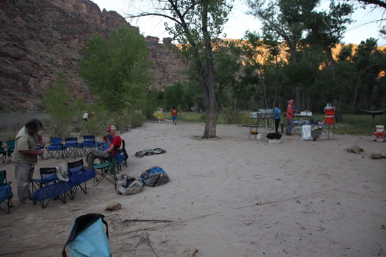 Campground scene with people setting up camp, blue chairs, and cooking equipment near a river.