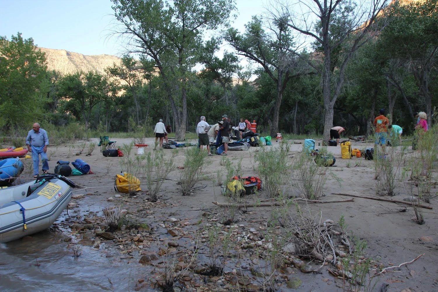 People gather on a sandy riverbank, surrounded by rafts, gear, and trees.