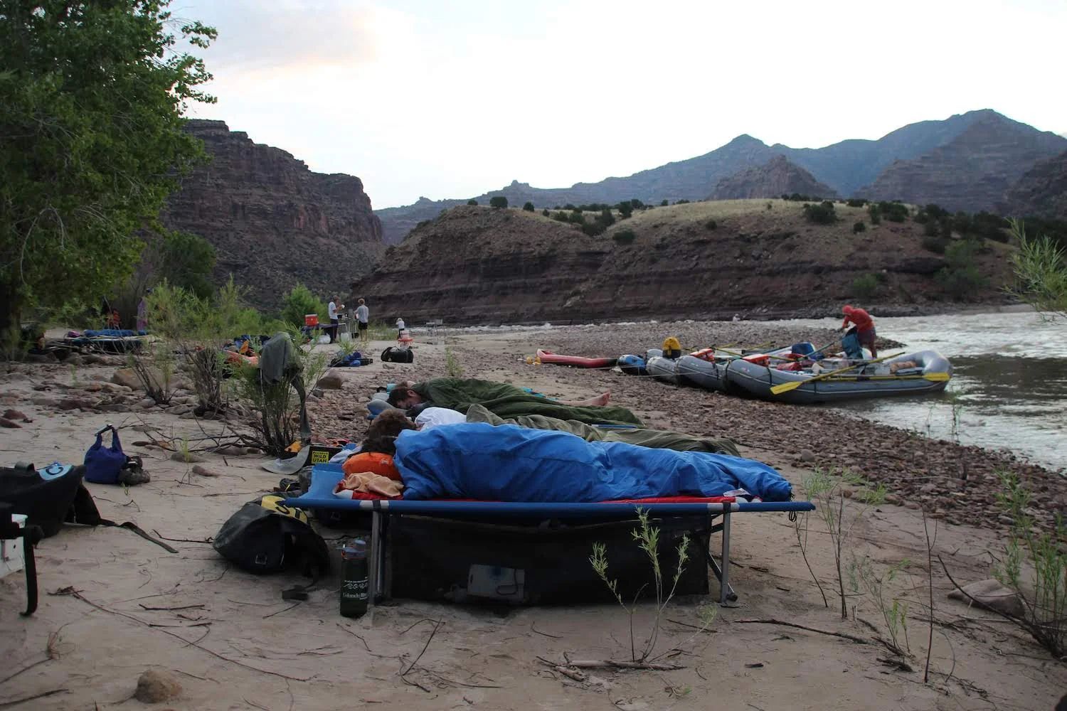 Camp site along a river. Rafts, gear, and people near a sandy shore with mountains in the background.