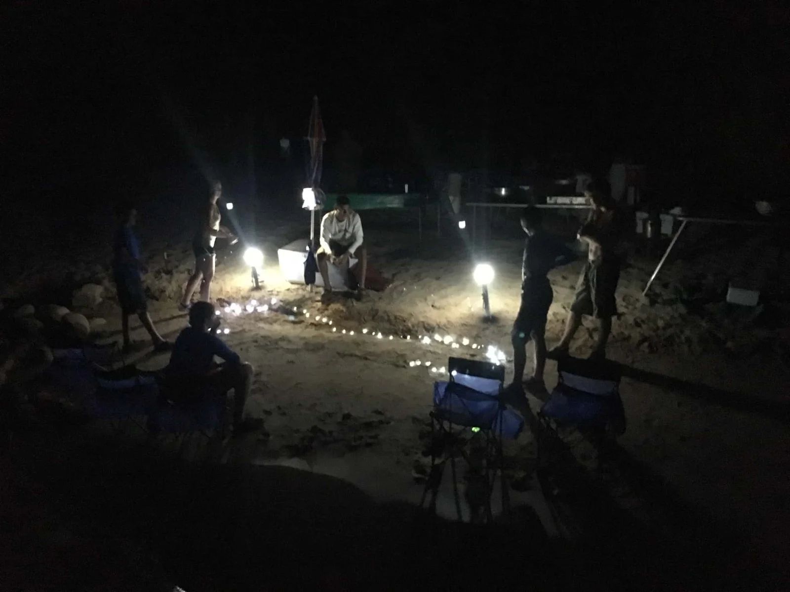 Group of people gathered outdoors at night, lit by string lights and lanterns, possibly camping or socializing.